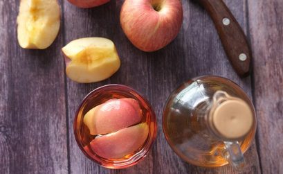 apple cider vinegar in a glass and decanter on a wooden table with whole and cut apples beside it