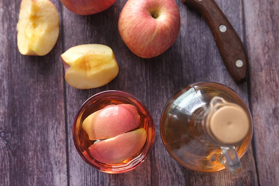 apple cider vinegar in a glass and decanter on a wooden table with whole and cut apples beside it